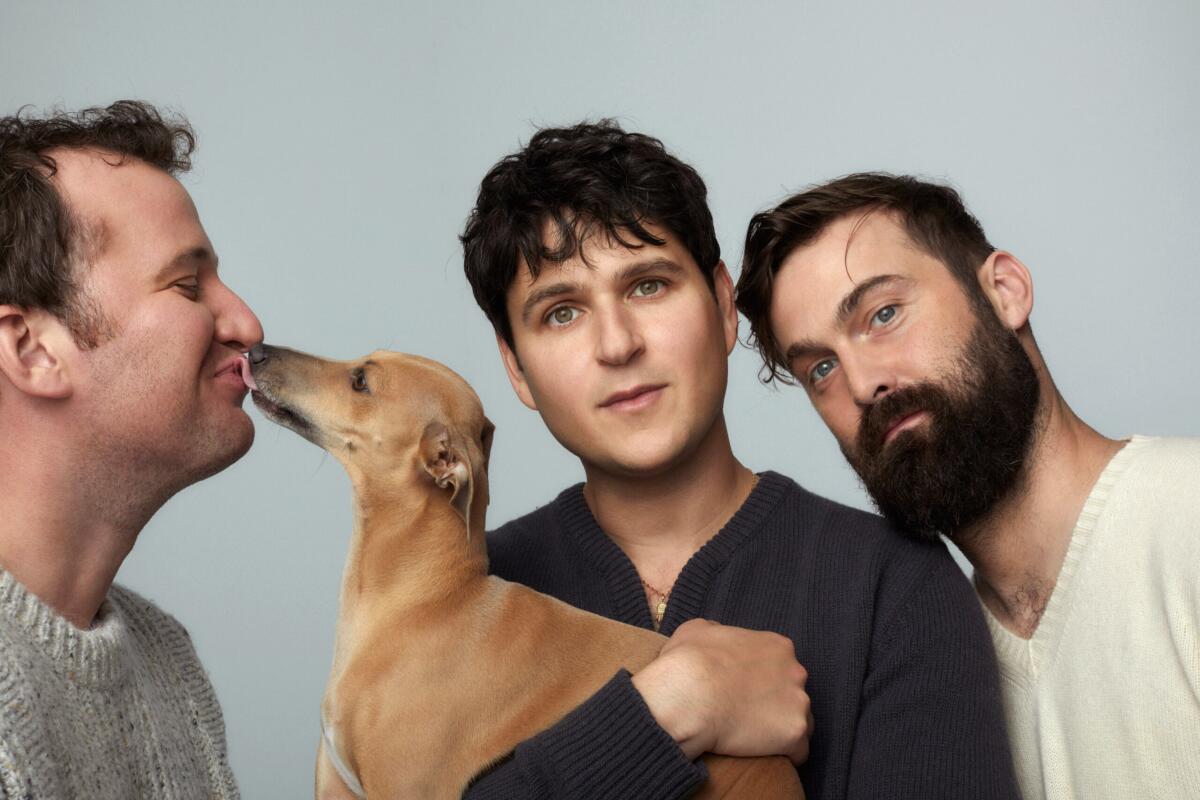Three male band members, one holding a dog that licks the nose of another.
