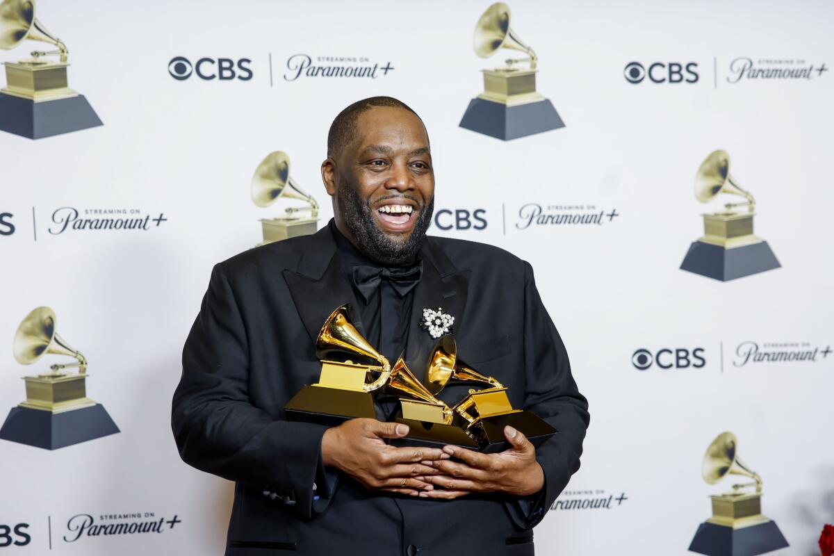 Killer Mike in a black suit holds 3 golden Grammy Awards in his arms as he smiles and poses in front of a white backdrop