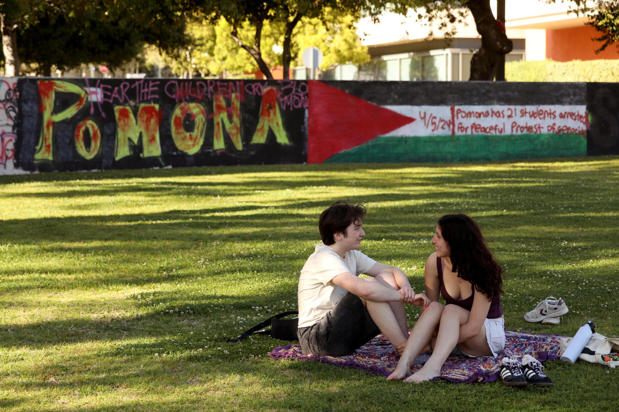 Pomona students Morgan Sydor and Rebecca Allen sit in a grassy area near a wall with writing on it.
