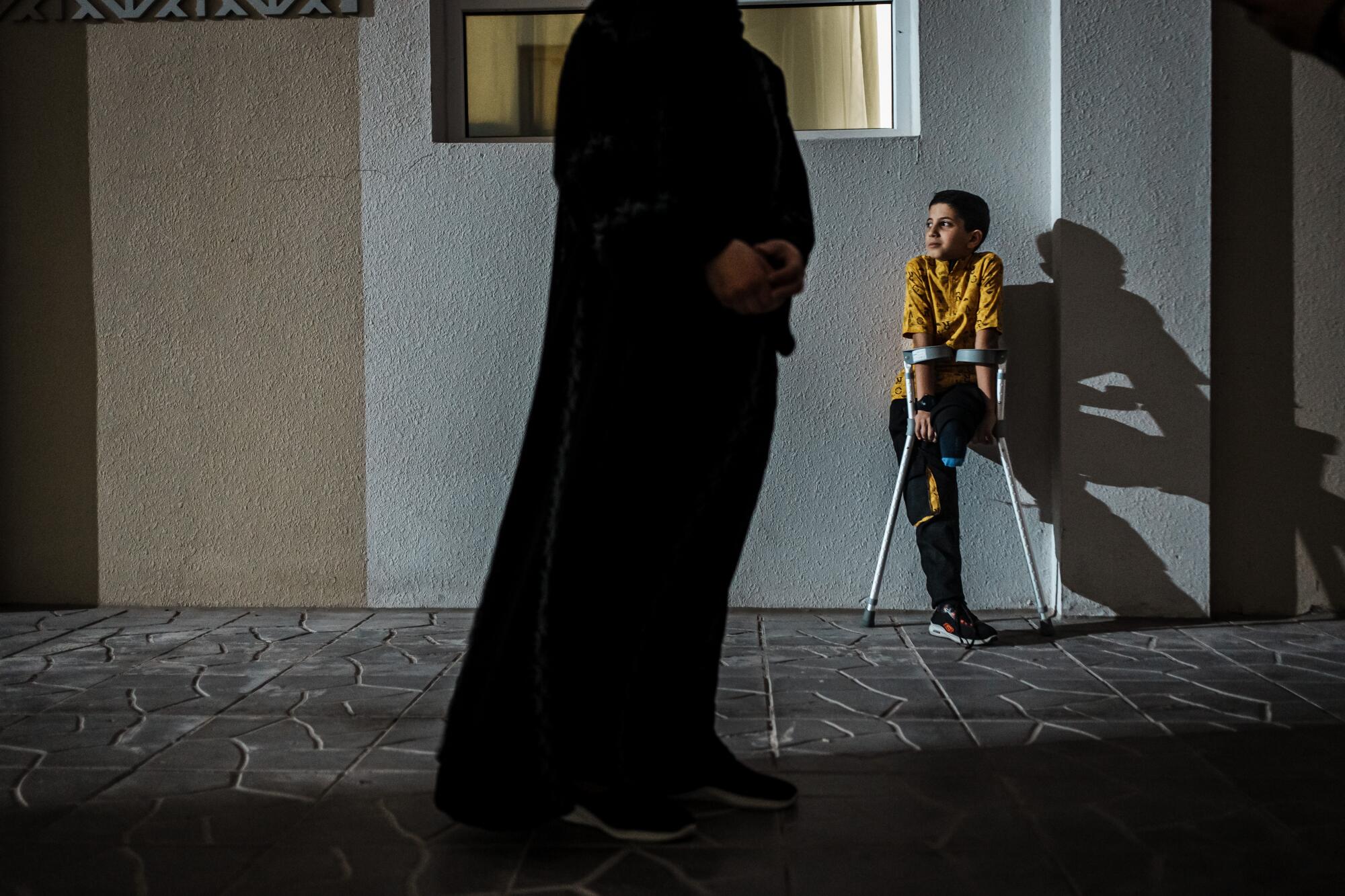一个单腿拄着拐杖的孩子，他的影子落在墙上，一个成年人站在前景中。