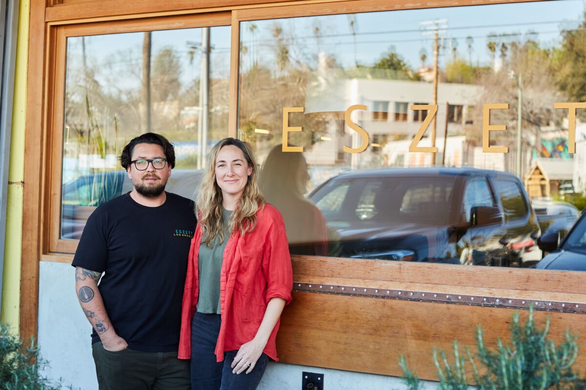 A man and a woman outside a mirrored restaurant window with the word "Eszett" painted on it.