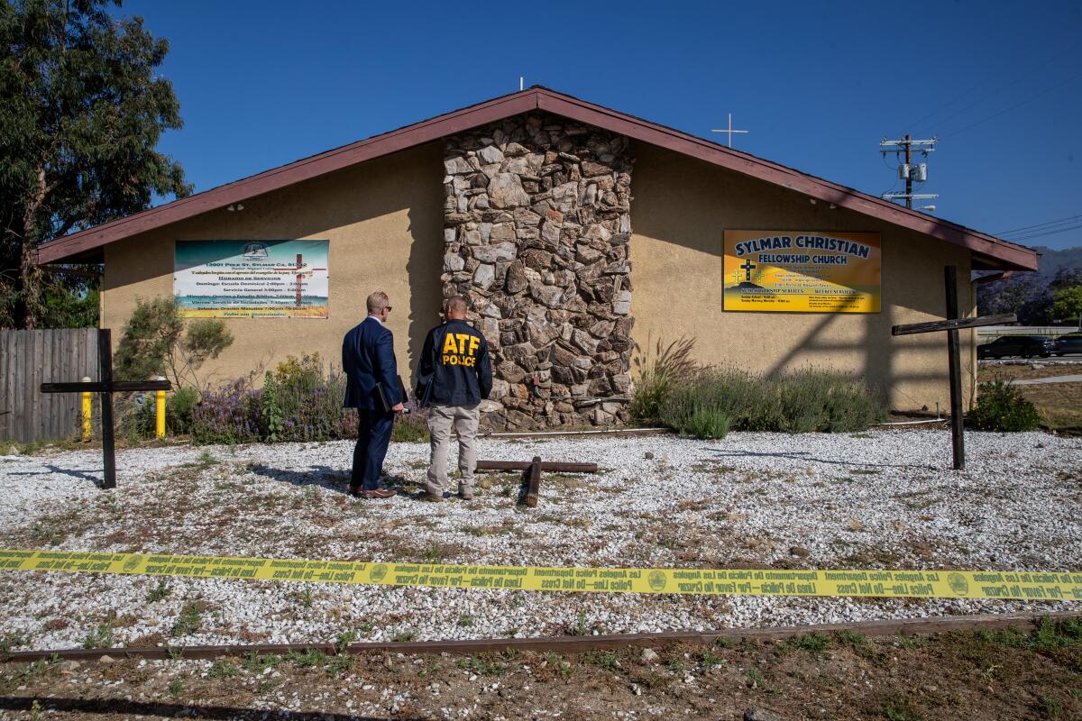 Two law enforcement officers stand next to burned wooden crosses behind crime scene tape outside a church building