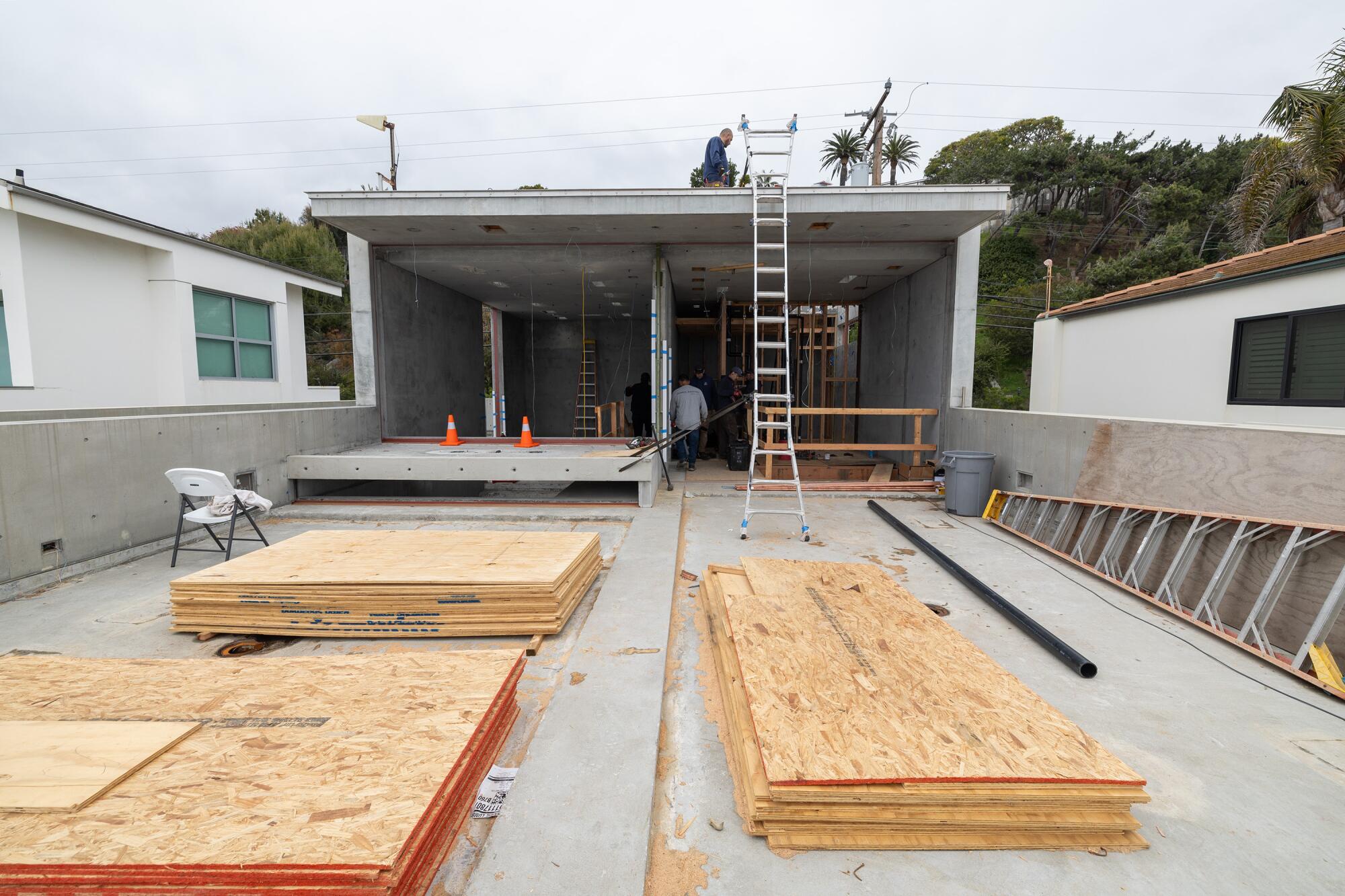 Stacks of plywood lie on a deck next to an open room. A ladder is propped against the building and a man is on the roof.