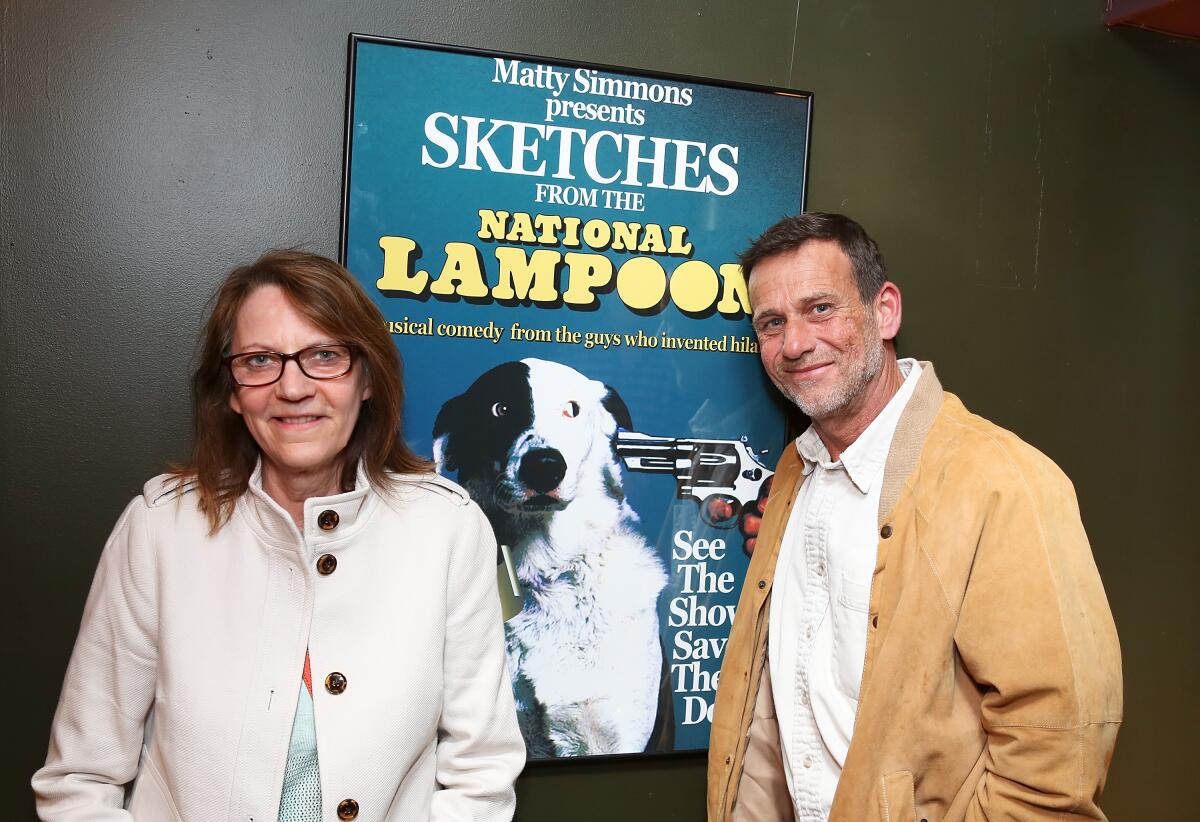 Judy Belushi Pisano and her nephew standing by a "Sketches From the National Lampoon" poster.