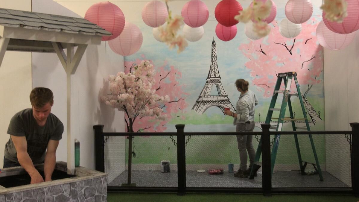 Muralist Hanna Daly paints a Paris scene as Ryan Erpelding, left, works on a wishing well Thursday for the opening later in the day of the touring pop-up exhibition "The Museum of What? Love Tour" in Encinitas.