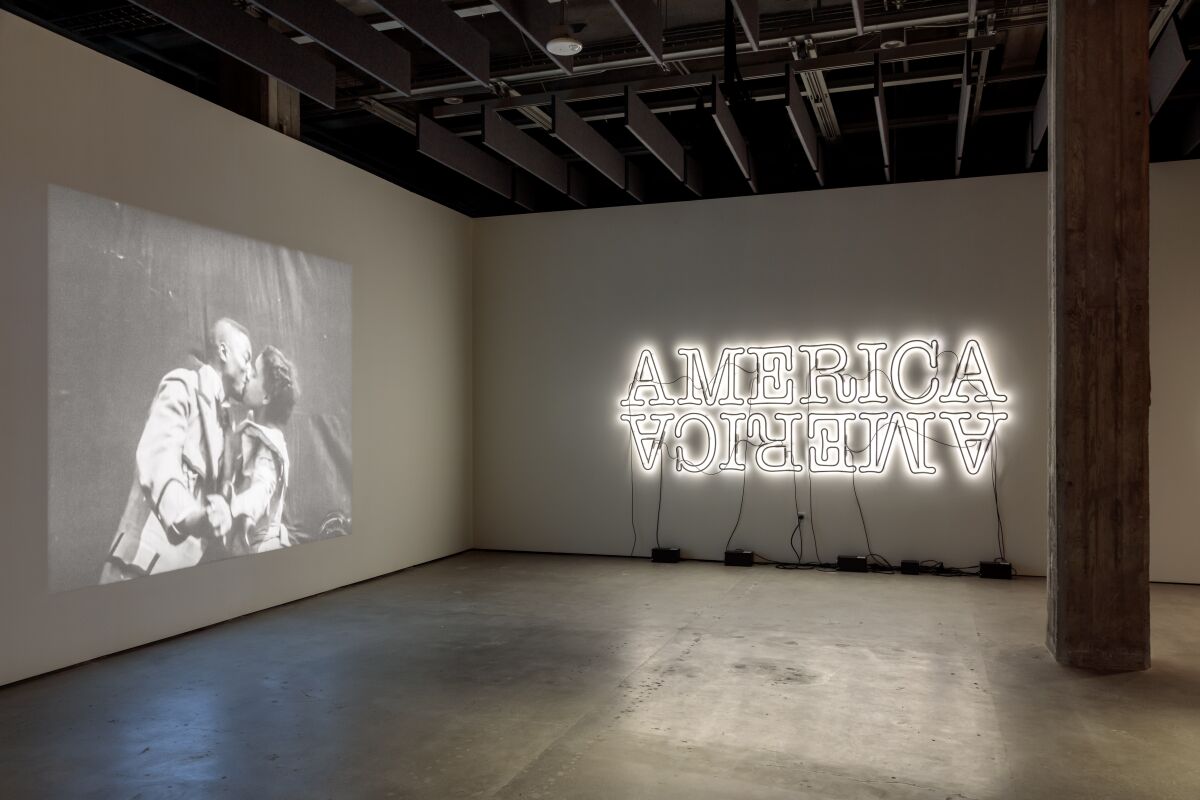 A view of a gallery shows a film of a Black couple kissing at left and a neon art work of the word "America."