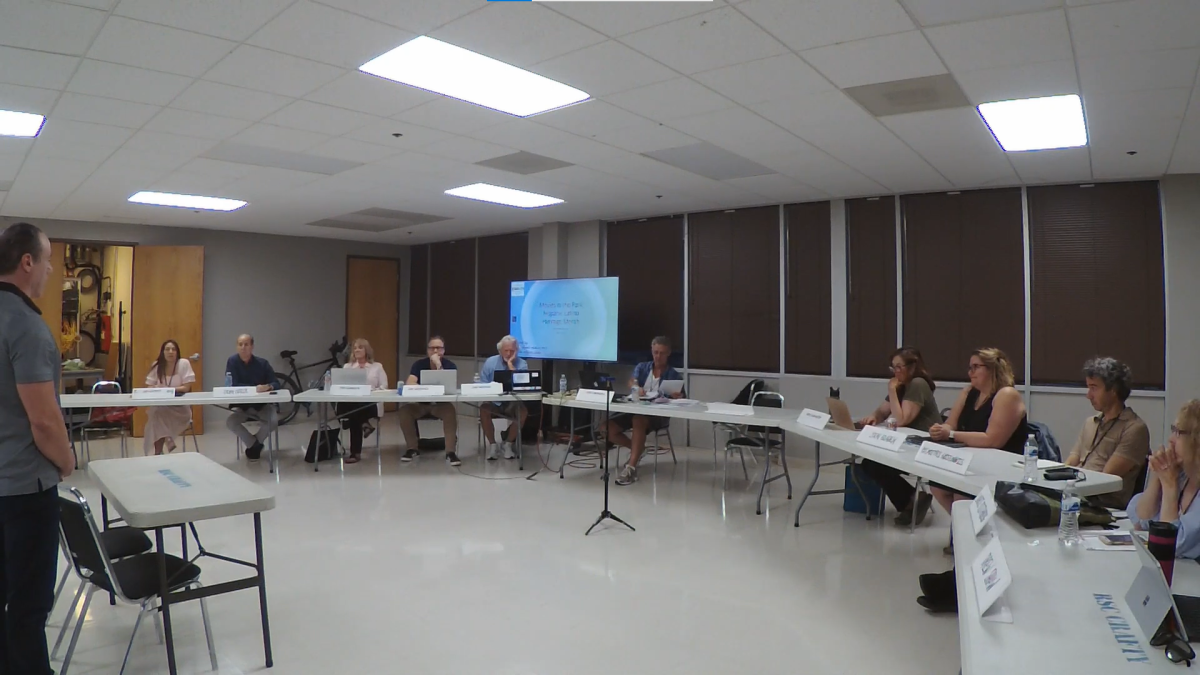 Screenshot of a public meeting in a room with people sitting at tables.