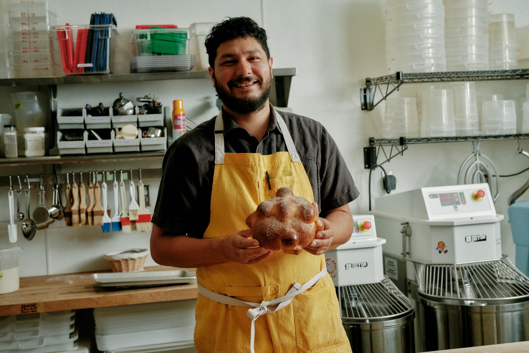 Gusto Bread owner Arturo Enciso, in a yellow apron, holds freshly baked pan de muerto.