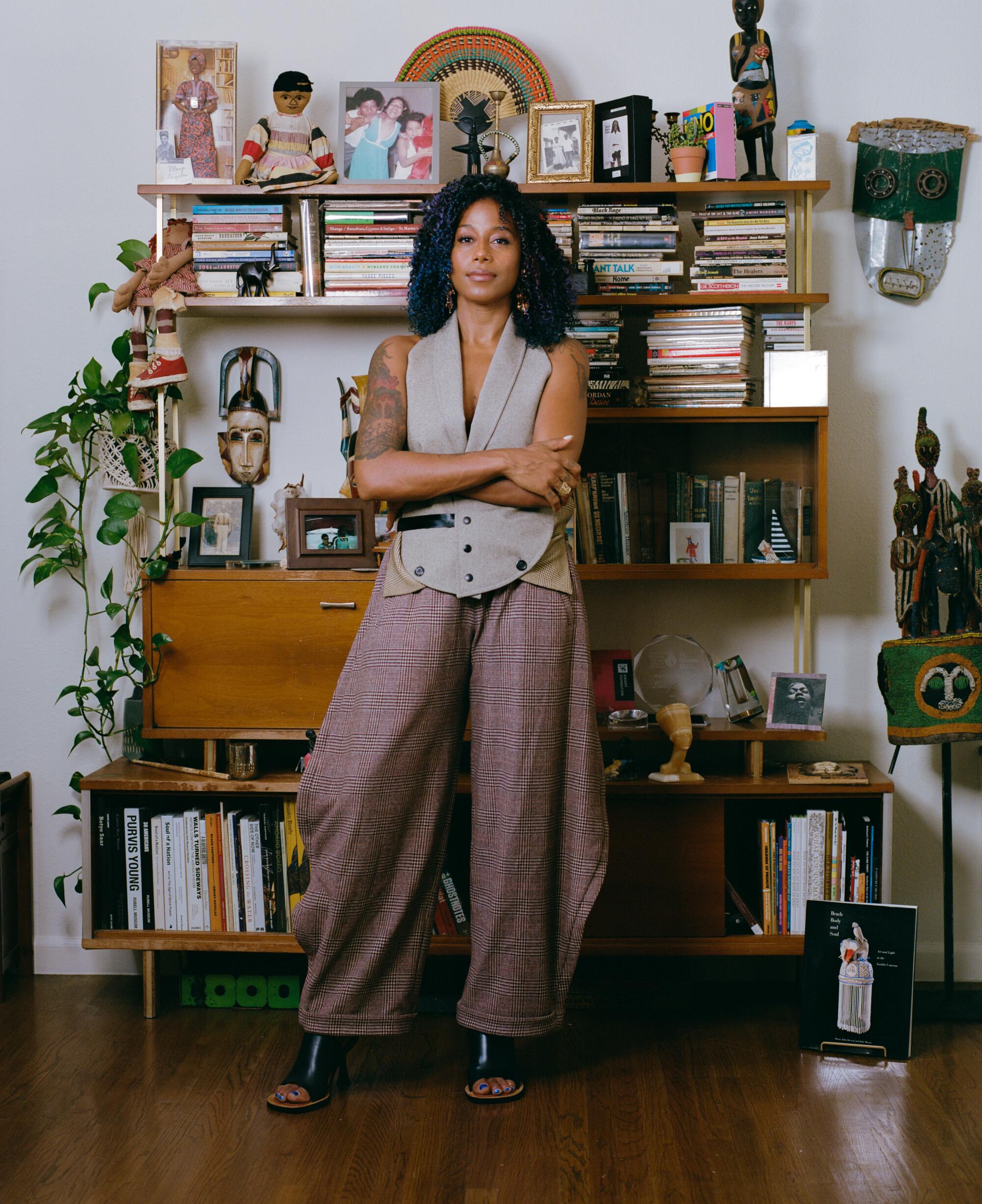 A woman stands, arms crossed, in front of a shelf filled with objects and books