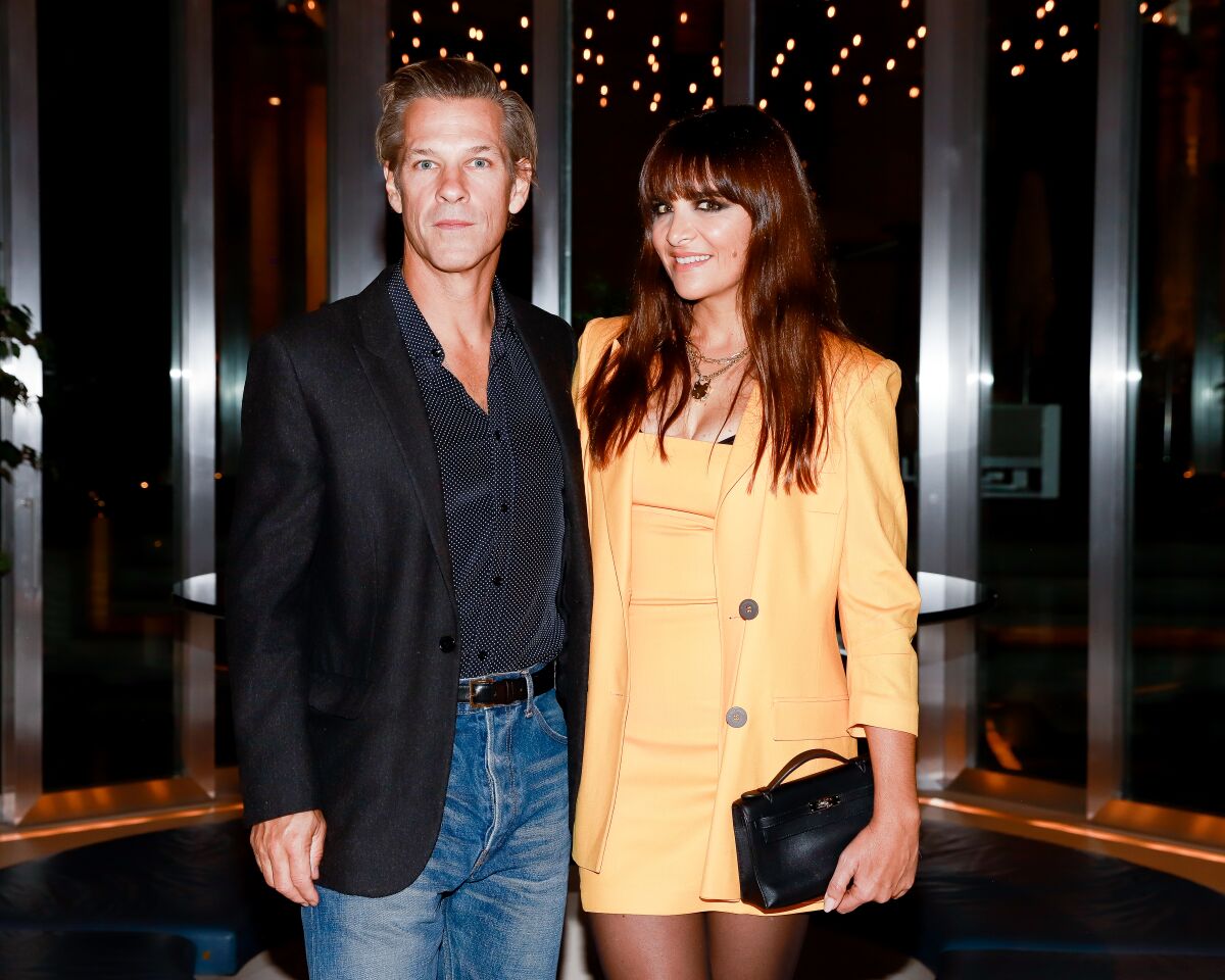 A man in dark shirt and blazer and blue jeans stands next to a woman in a short peach dress and blazer.