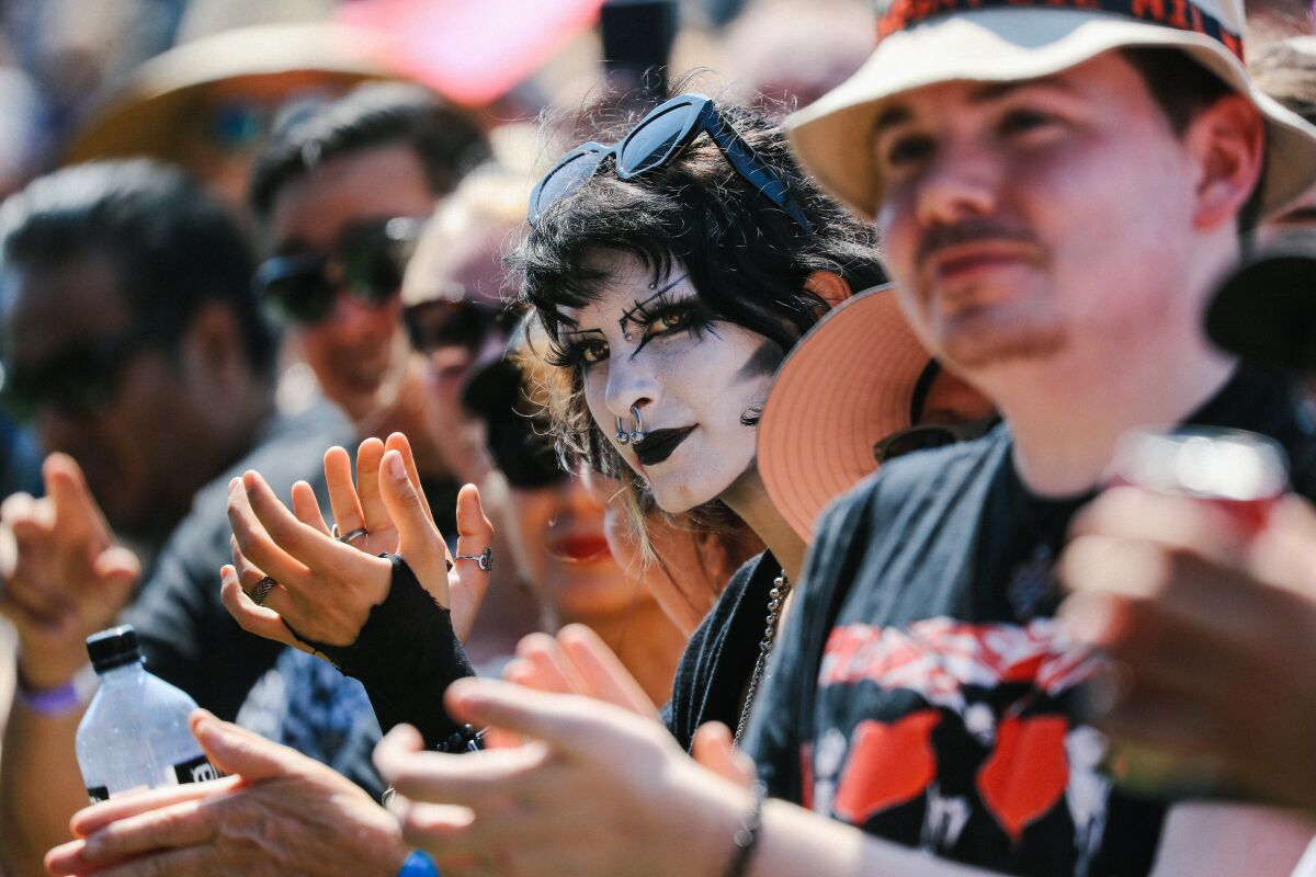 A person with a white face paint and exaggerated makeup in an outdoor crowd, all applauding.