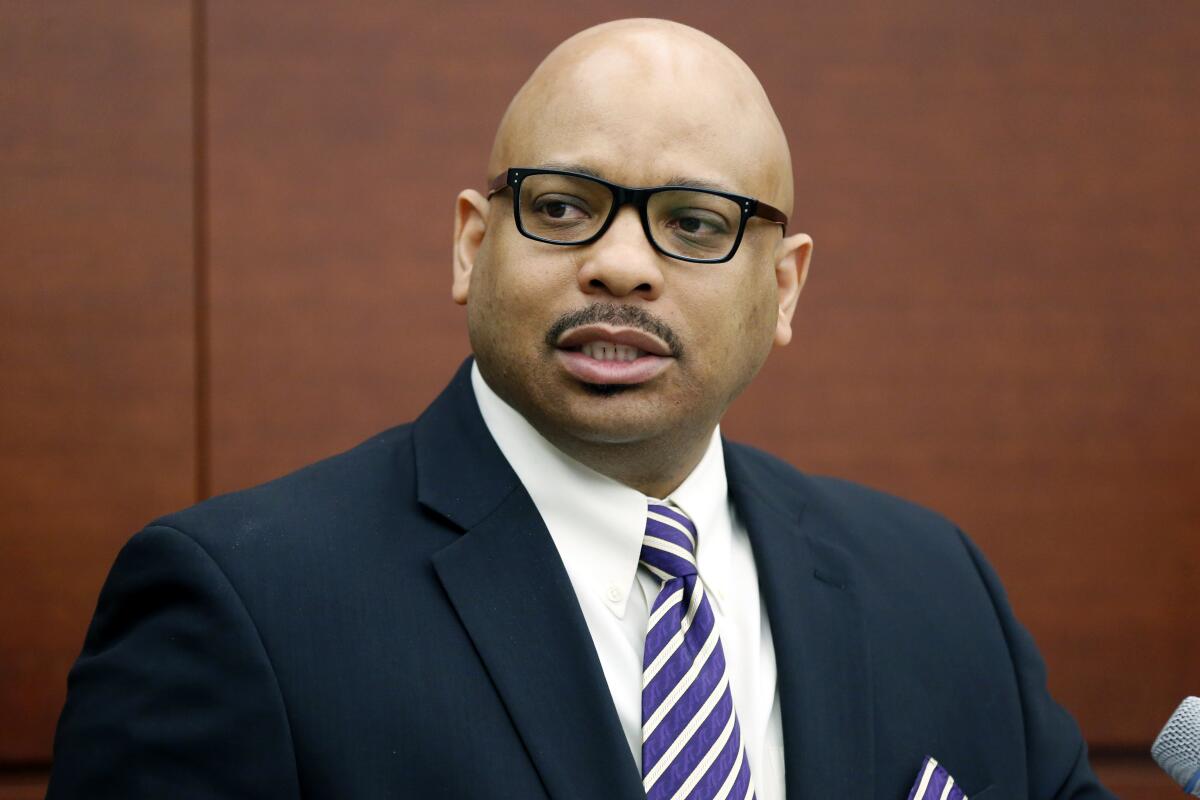 A bald man with a dark mustache, in a dark suit and purple striped tie, speaks