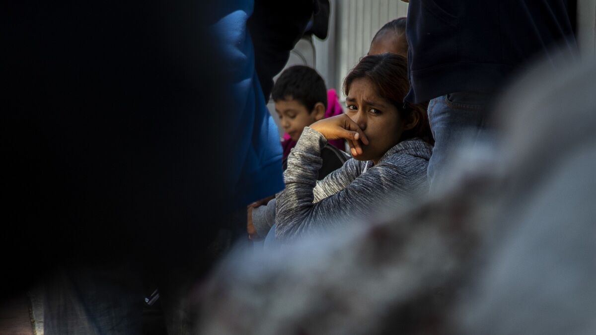 An immigrant girl sits with her family members at the border while they track down the keeper of the notebook.
