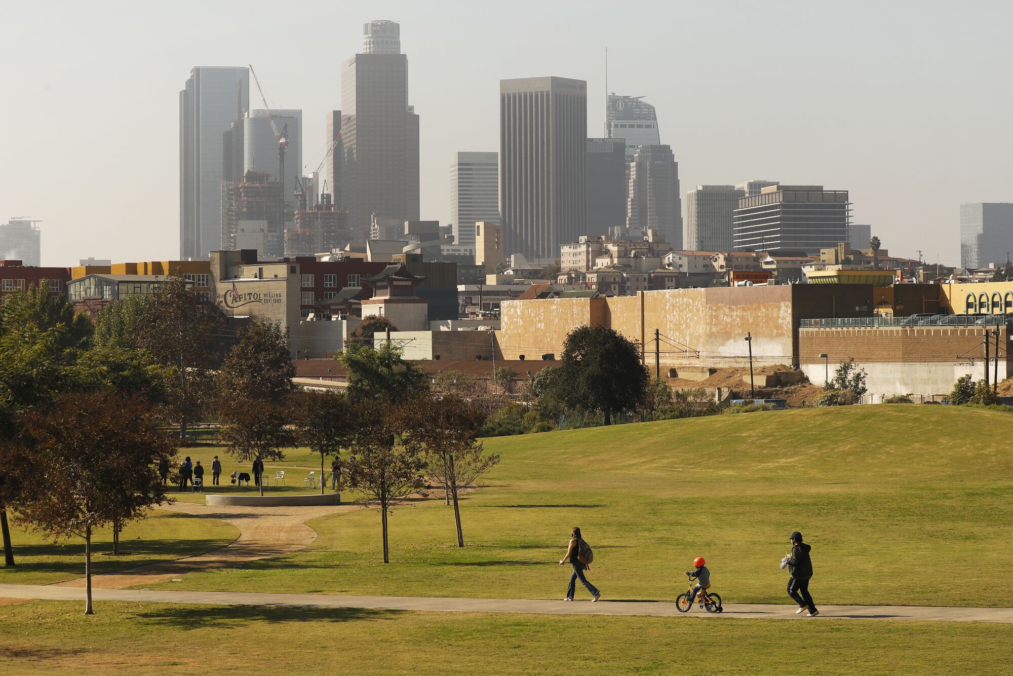 People walk and bike on a path through Los Angeles State Historic Park, with the downtown skyline in the background