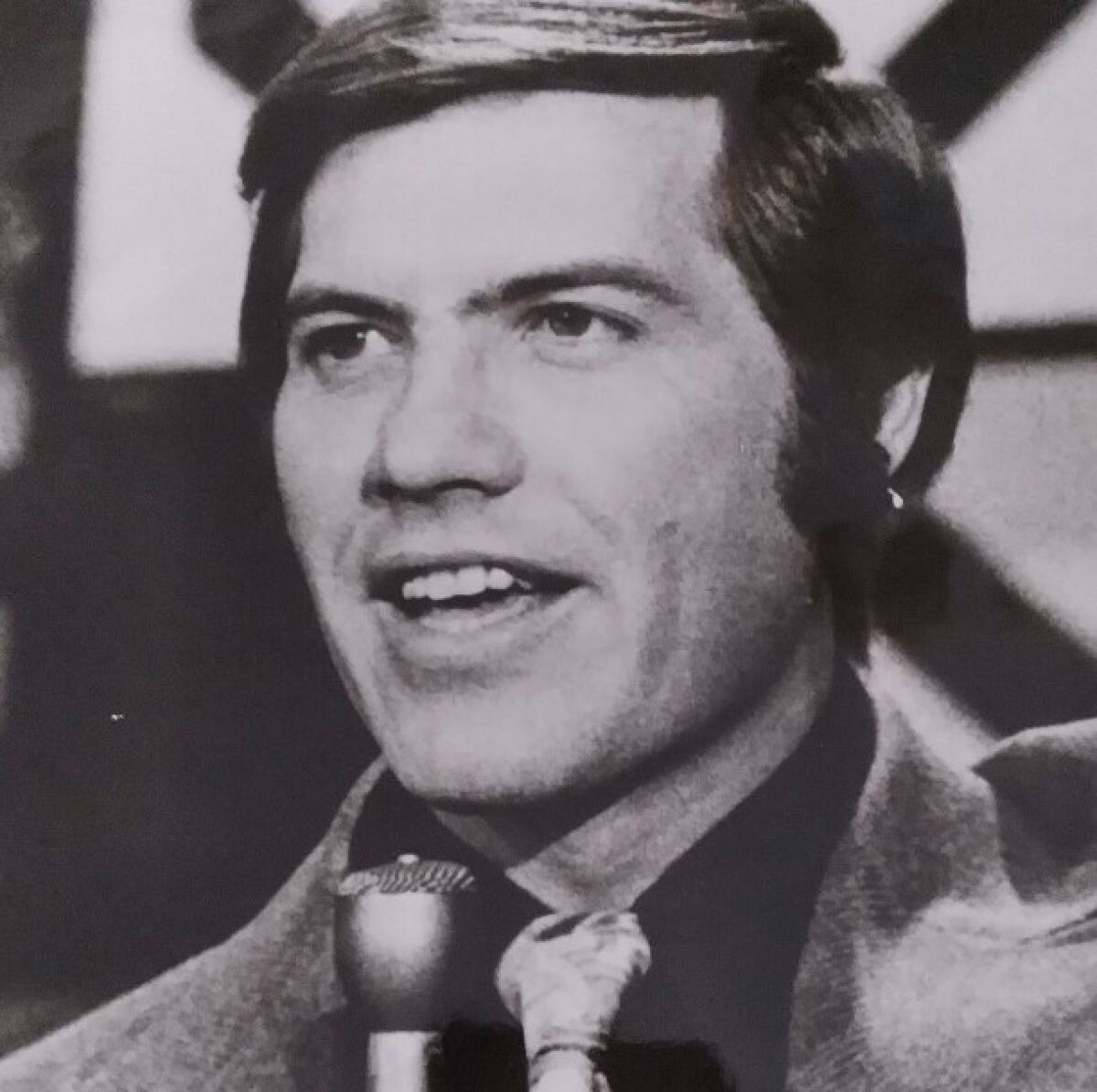 A close-up of Sam Riddle in a suit, holding a microphone.