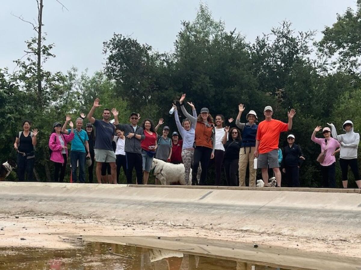 A group of about 20 people, by a pond on a trail, wave.