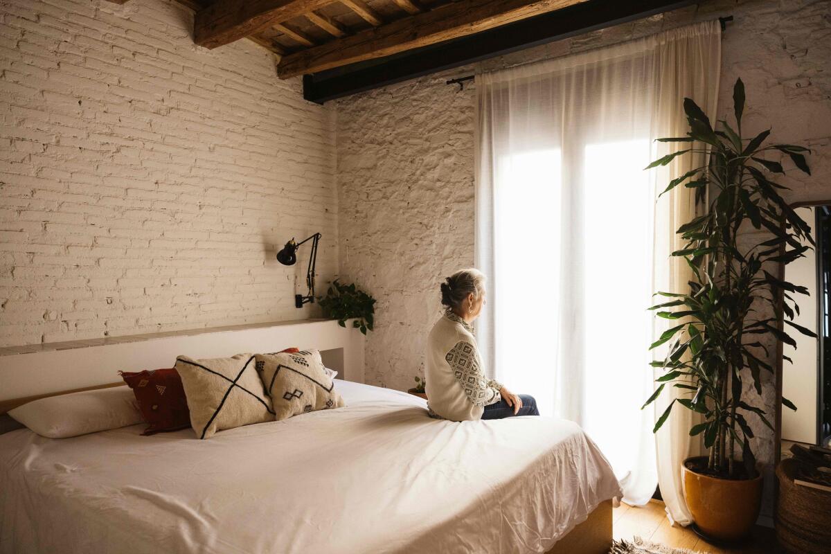 Portrait of thoughtful real mature senior woman sitting on bed in front of a window in a bedroom.