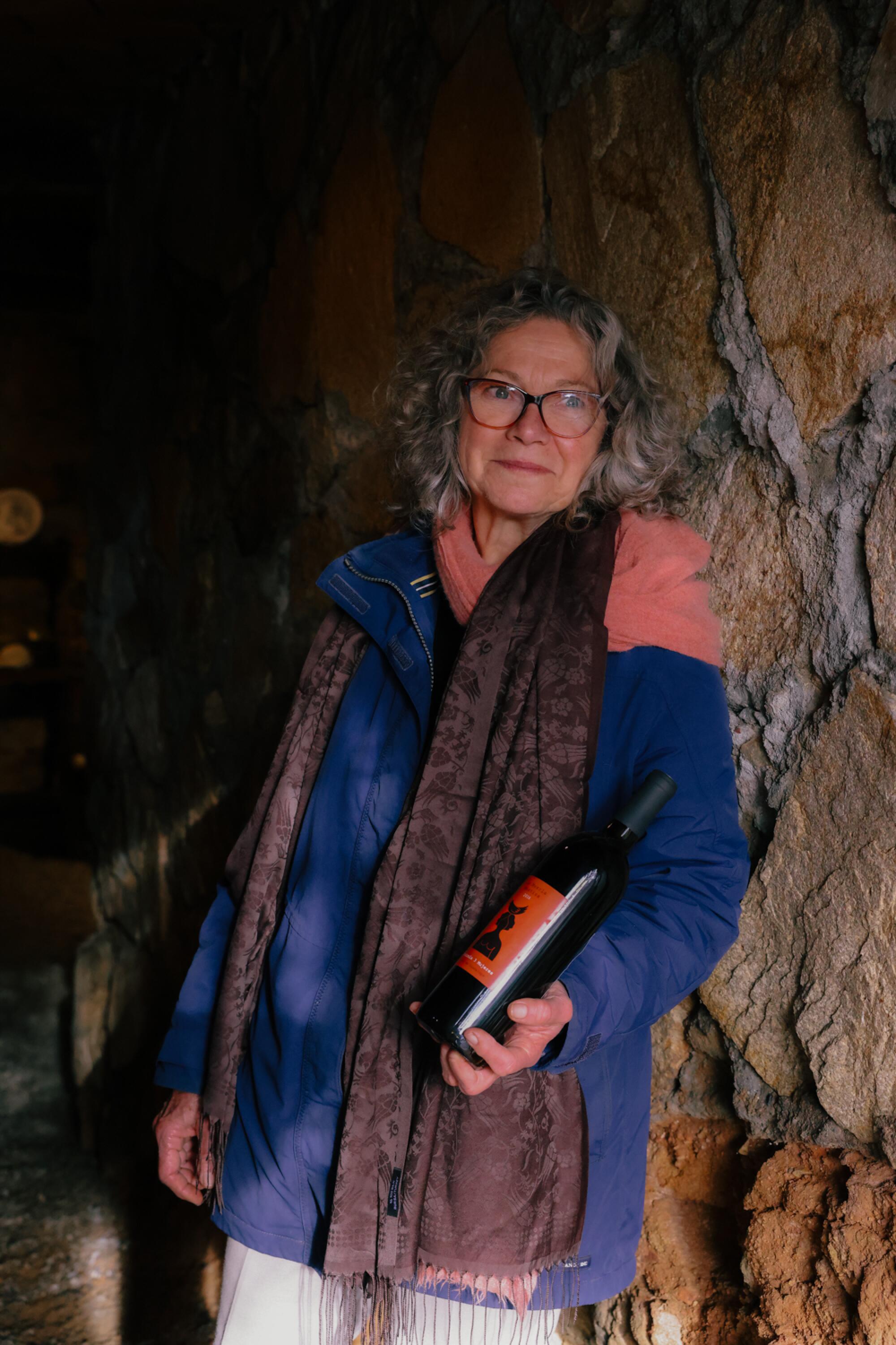 A woman leans against an uneven stone wall, holding a bottle of wine