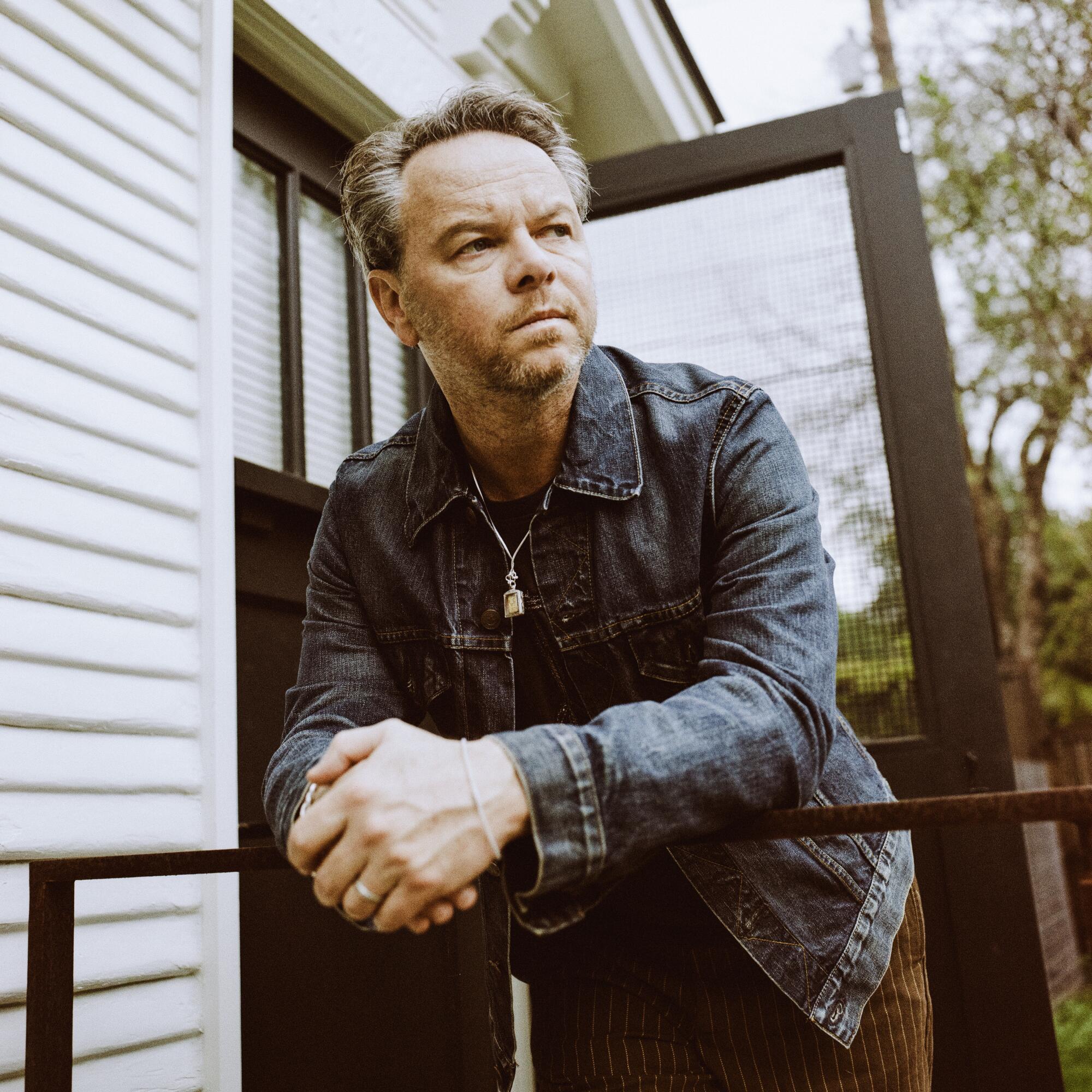 Noah Hawley, in a denim jacket and brown pants, leans against the railing of a patio.