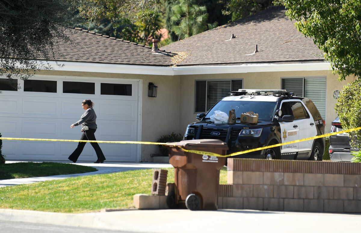 A person walks in front of the garage of a one-story house while a police car is parked in the driveway.