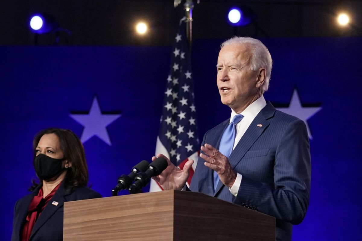 Joe Biden gestures with both hands as he speaks at a lectern, with Kamala Harris standing nearby in a mask