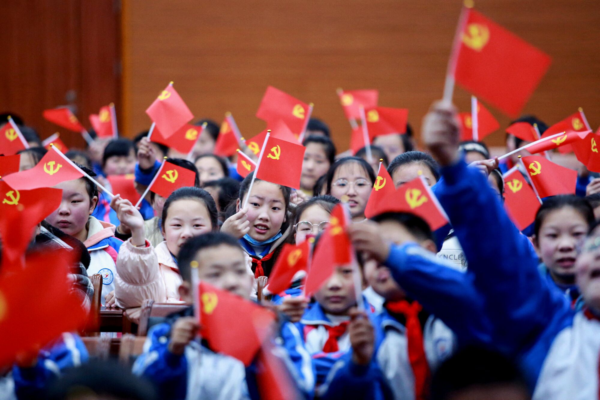 A big group of children wave red flags bearing yellow hammer and sickle symbol in corner