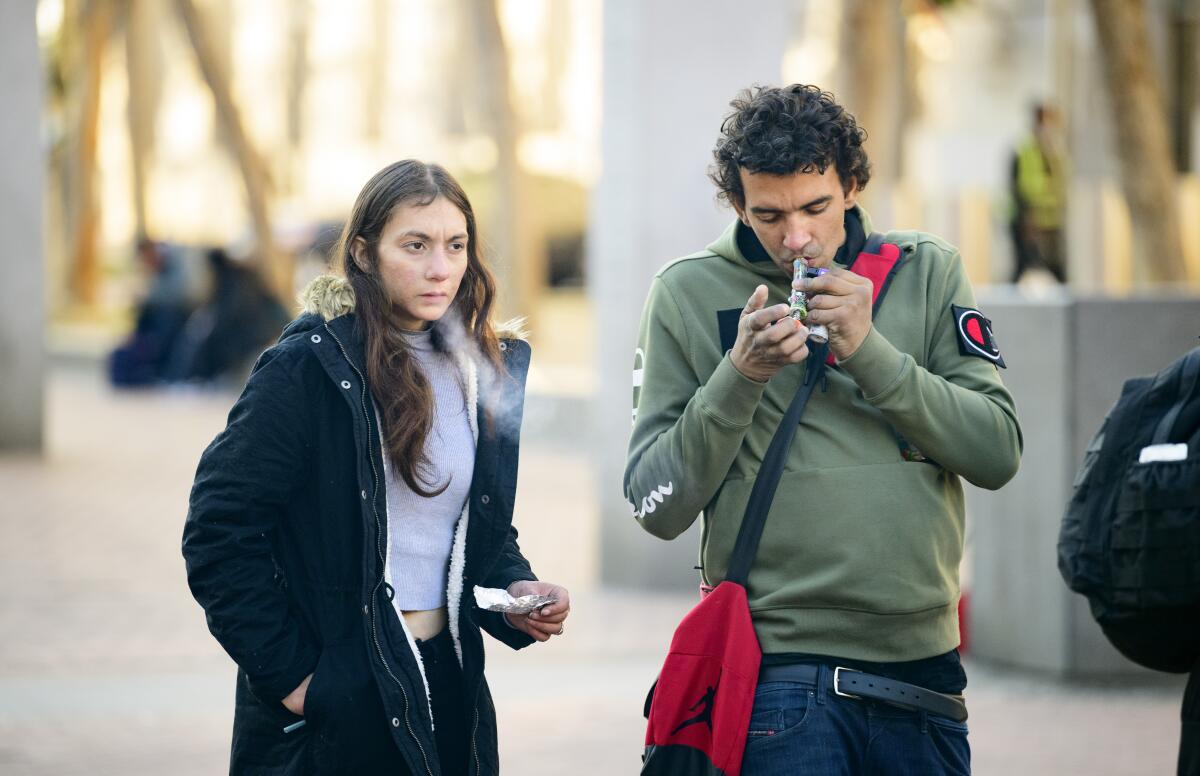 Two people stand outdoors. One smokes through a small pipe, and the other exhales a stream of smoke.