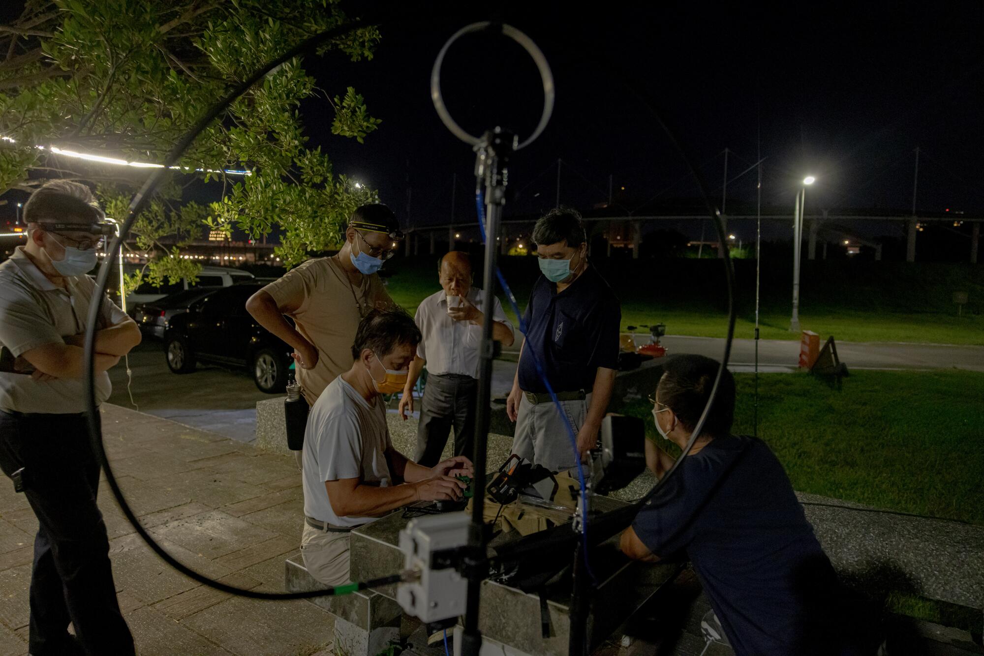 Several men gather around two people seated at a table near a pole with a large circular cable 