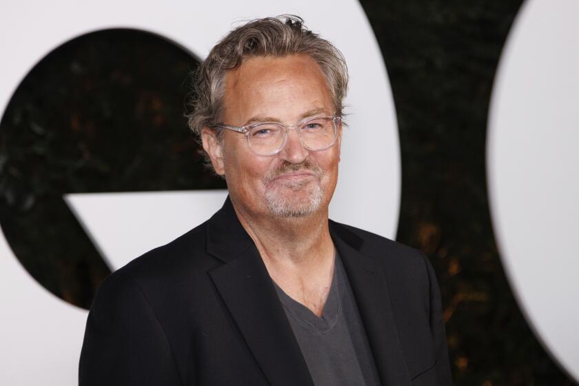 Matthew Perry smiles with his mouth closed while wearing glasses a gray shirt and a black suit jacket