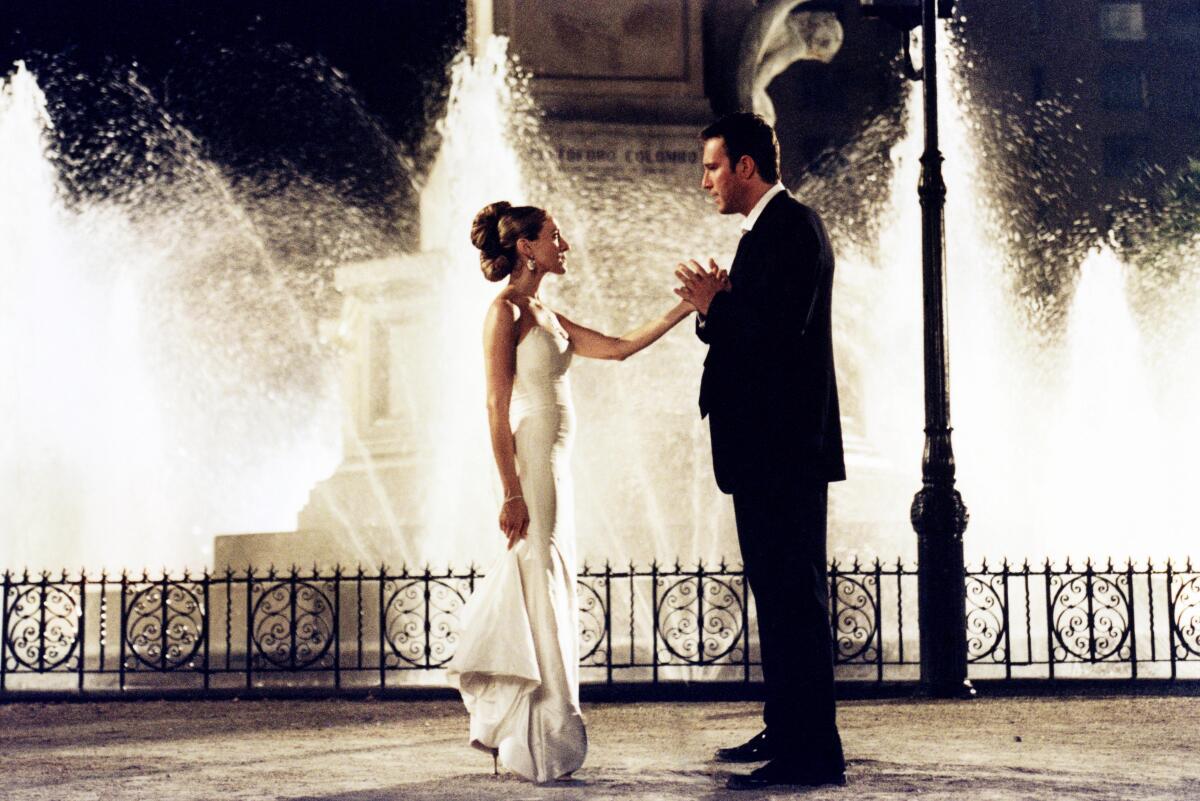 Before a water fountain, Carrie, in a white dress, stands with her arm stretched outward to Aidan, who is in a black suit.