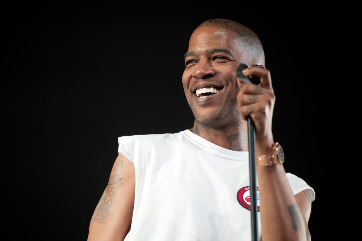 Kid Cudi smiling onstage and holding a mic in its stand while clad in a white crewneck with cutoff sleeves and a red logo