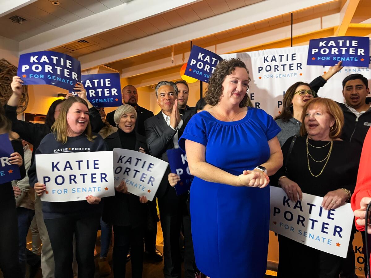 Katie Porter looks ahead as several supporters, many holding blue or white signs for her Senate campaign, cheer by her side