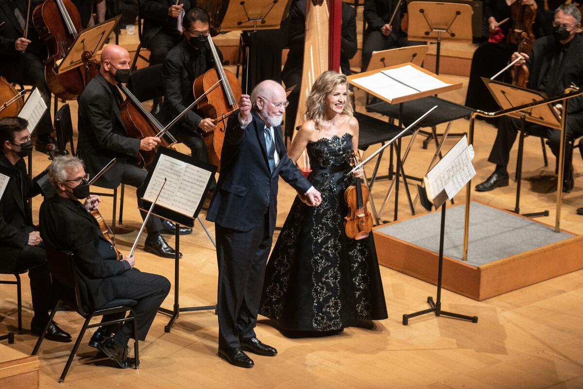 A man with a baton and a woman holding a violin stand before an orchestra and acknowledge the audience. 