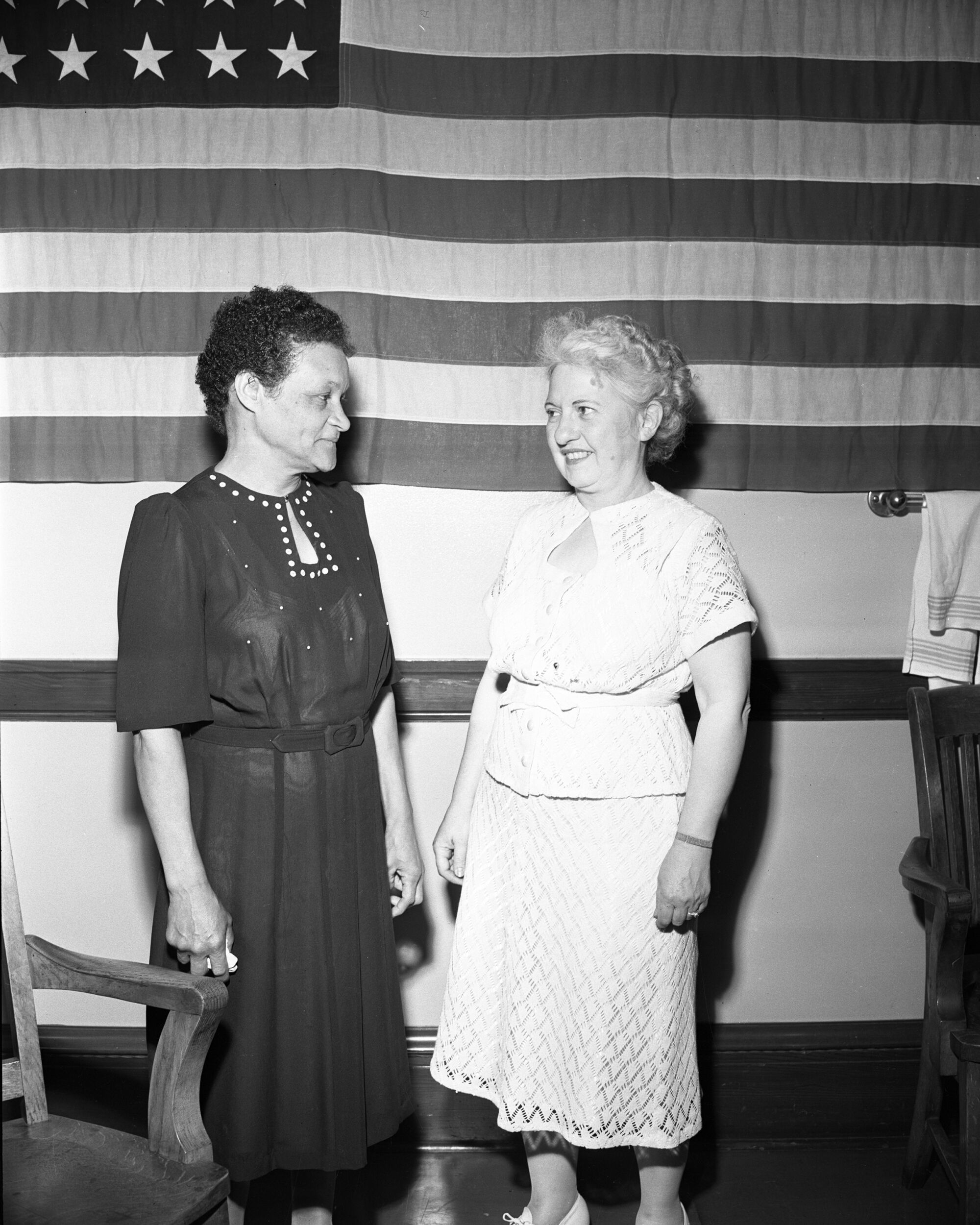 Dora Jones and Betty Marshal Graydon smile at each other in front of a U.S. flag