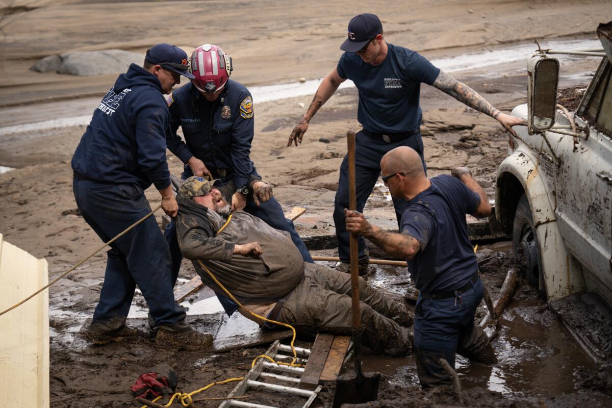 A man in soiled clothes wincing as four rescuers, one knee-deep in mud, free him from quicksand with rope