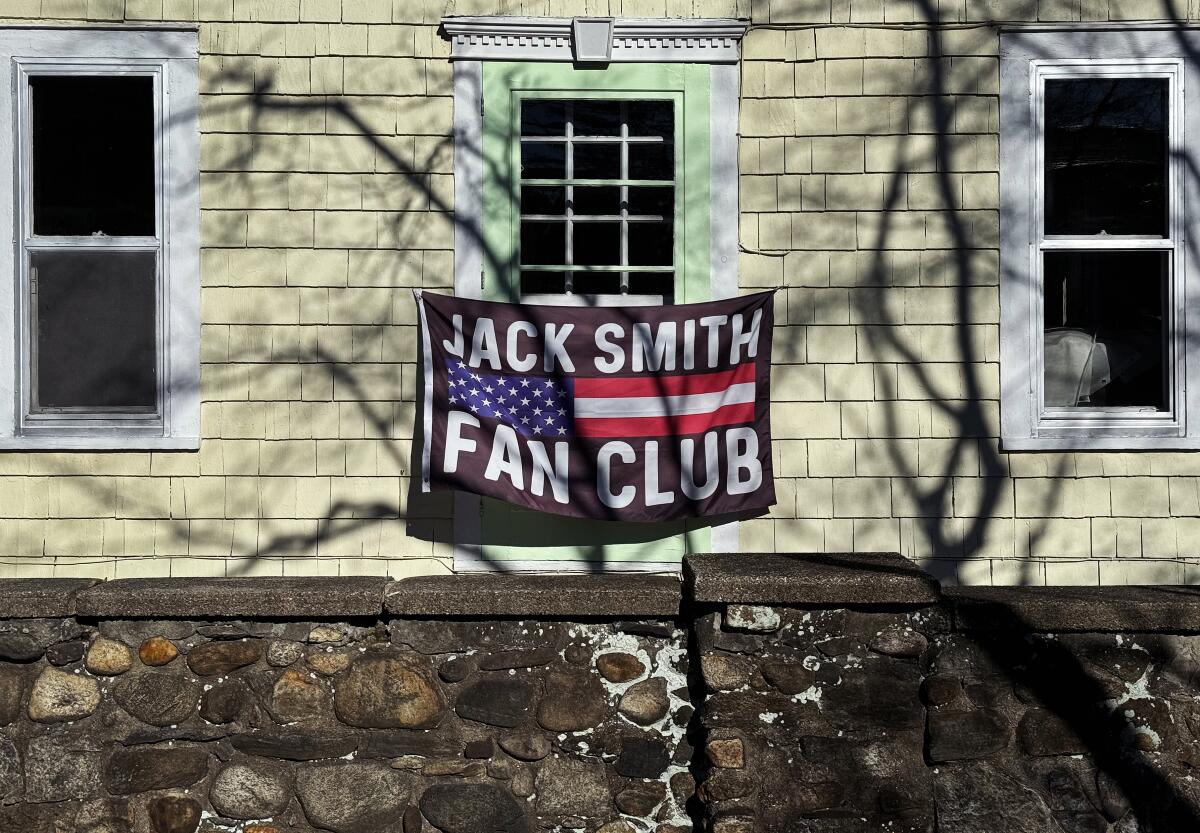 A flag that reads "Jack Smith Fan Club" hung above a stone foundation on a shingled house 