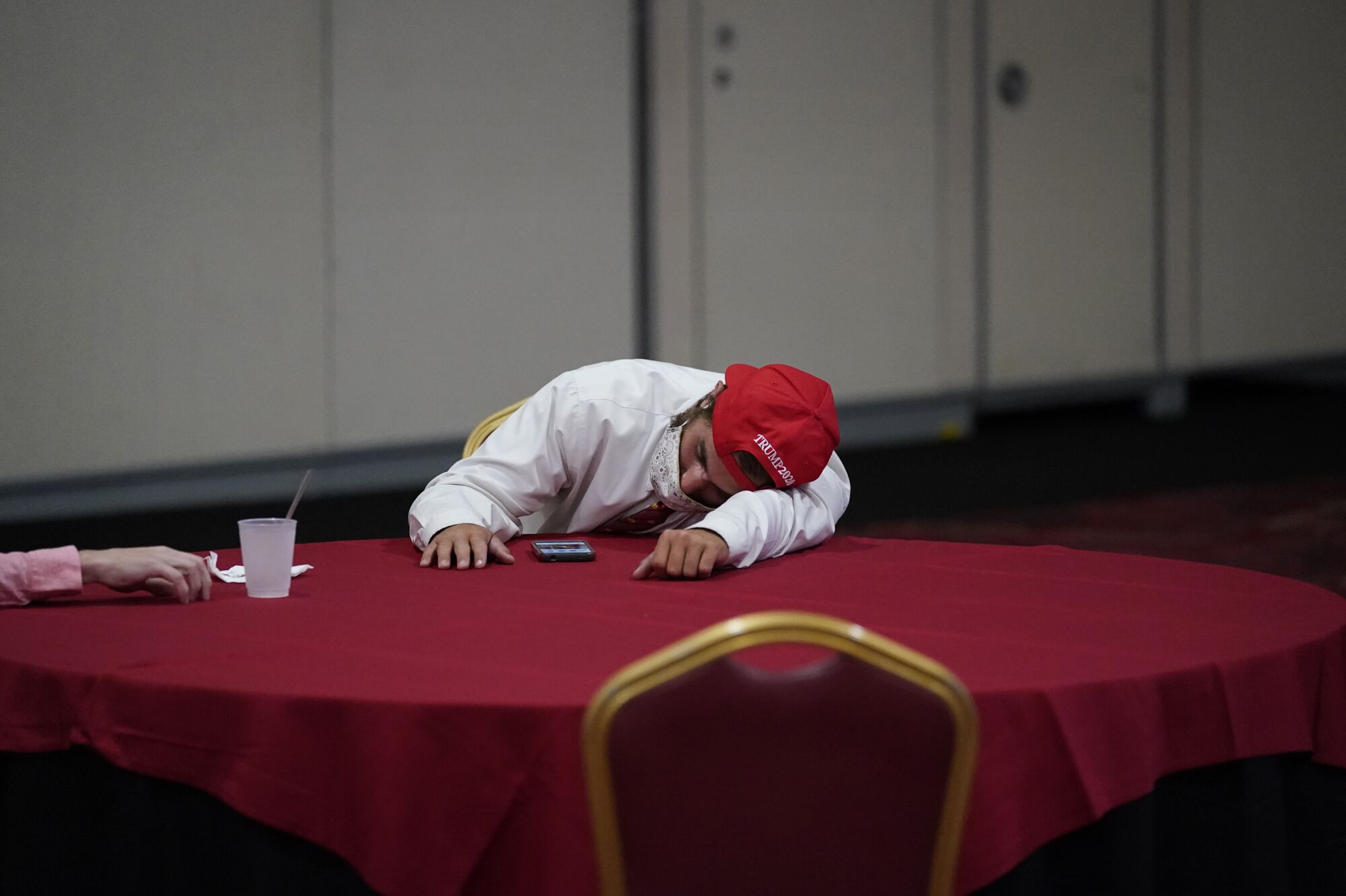 A man in a backwards red Trump 2020 hat rests on a table
