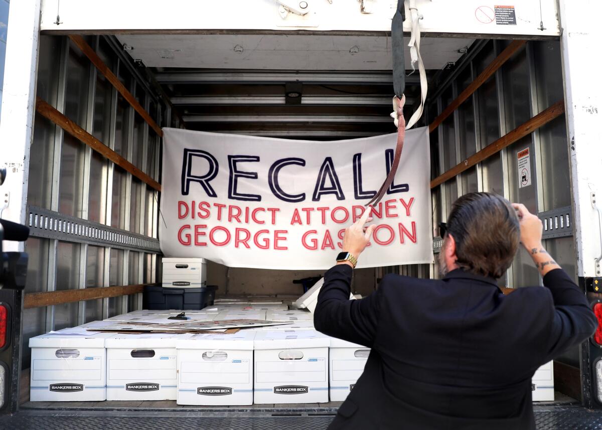 A man, facing away, opens a back of a delivery truck where signs hang reading "Recall District Attorney George Gascon."