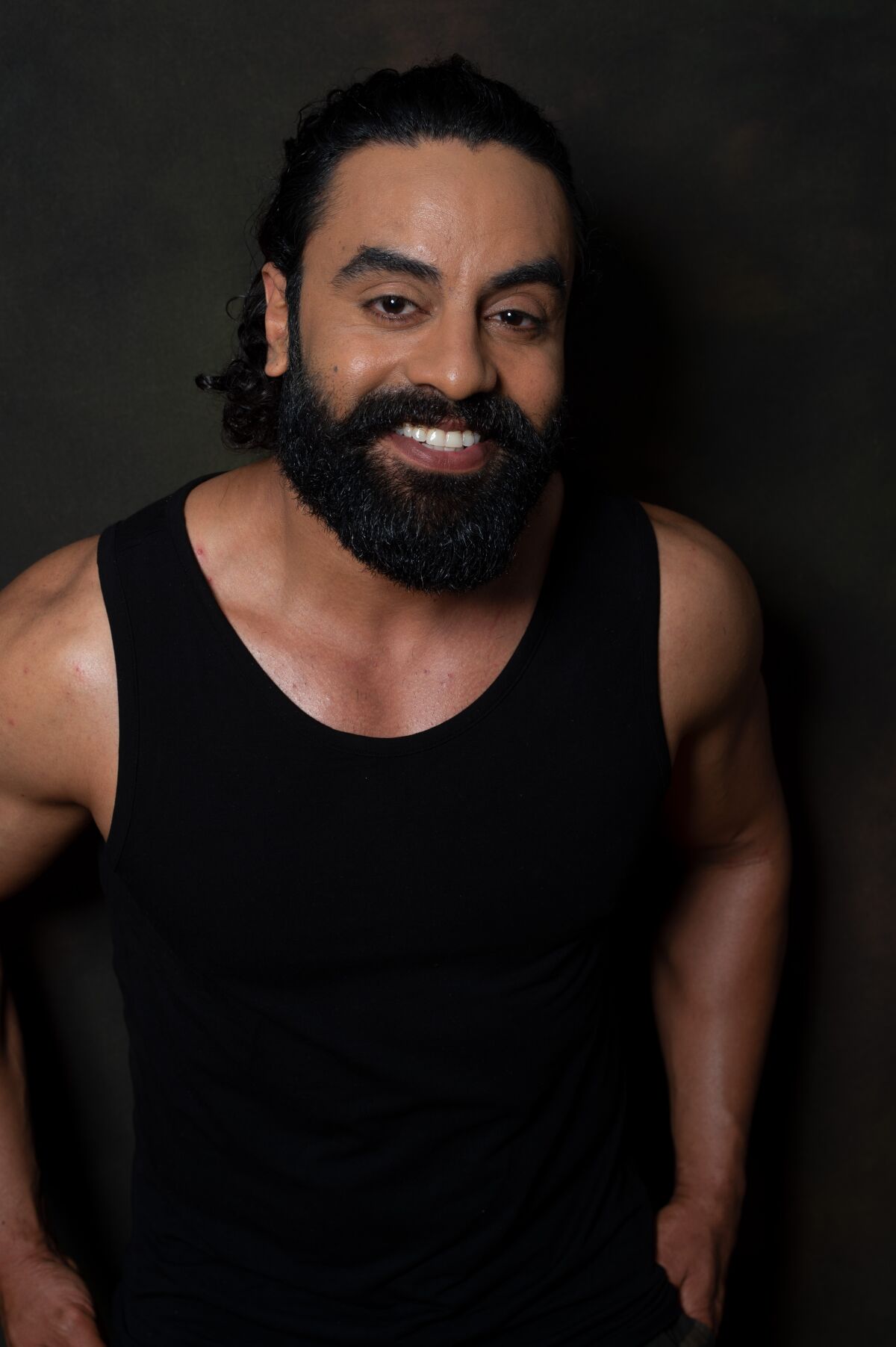 A man with a thick black beard in a black tank top smiles with his hands at his sides