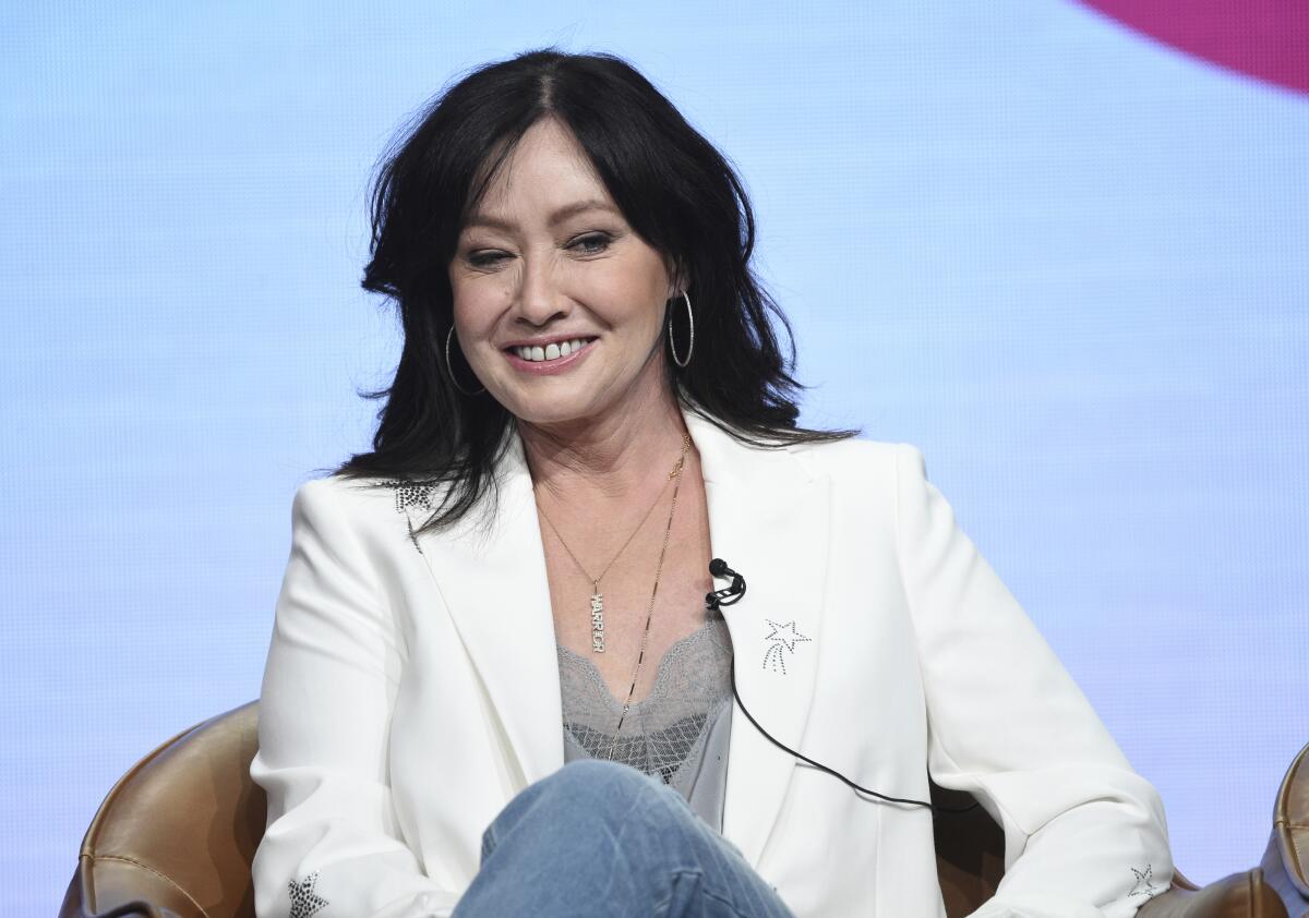 Shannon Doherty in a white blazer, jeans and a gray shirt sitting on a chair with her legs crossed