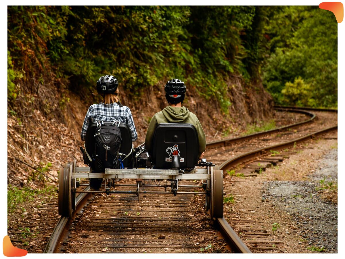 Two people in bike helmets pedal a low vehicle on railroad tracks.