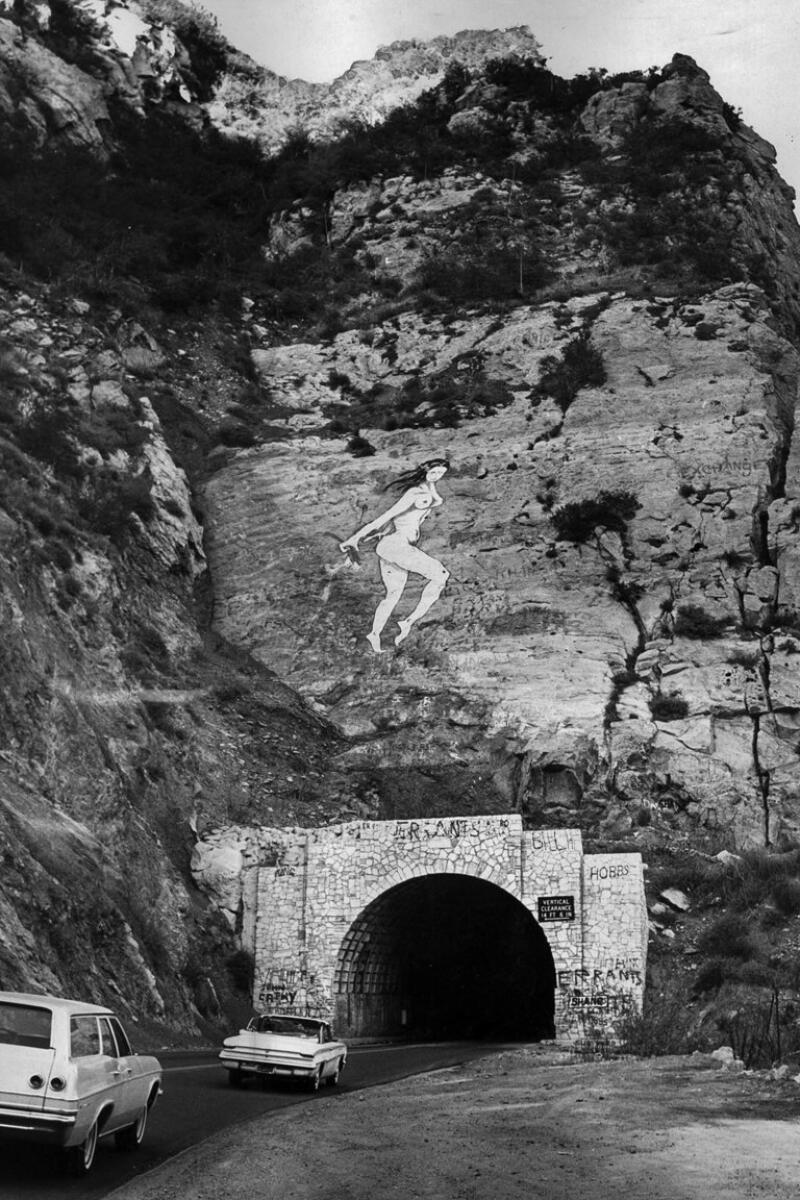 Black and white photo shows cars driving into the Malibu Canyon Road tunnel. A mural of a nude woman floats above the tunnel entrance. 