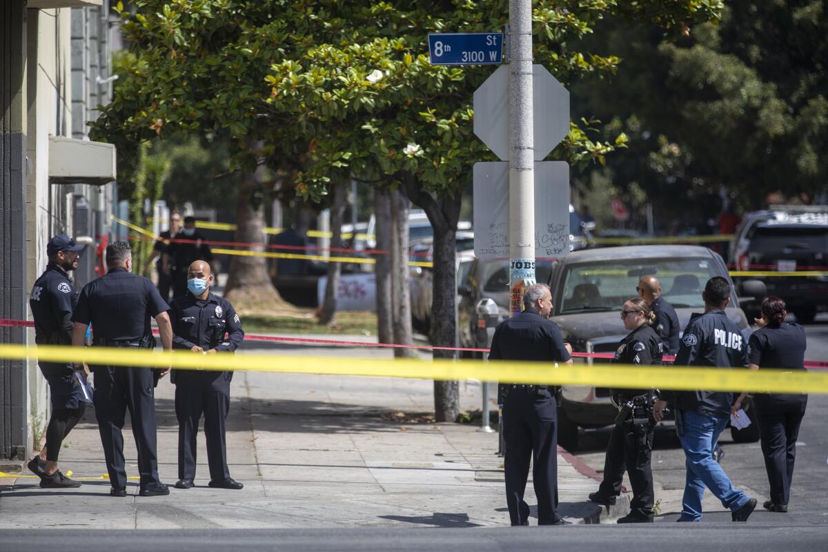 LAPD officers stand on a sidewalk behind yellow police tape.