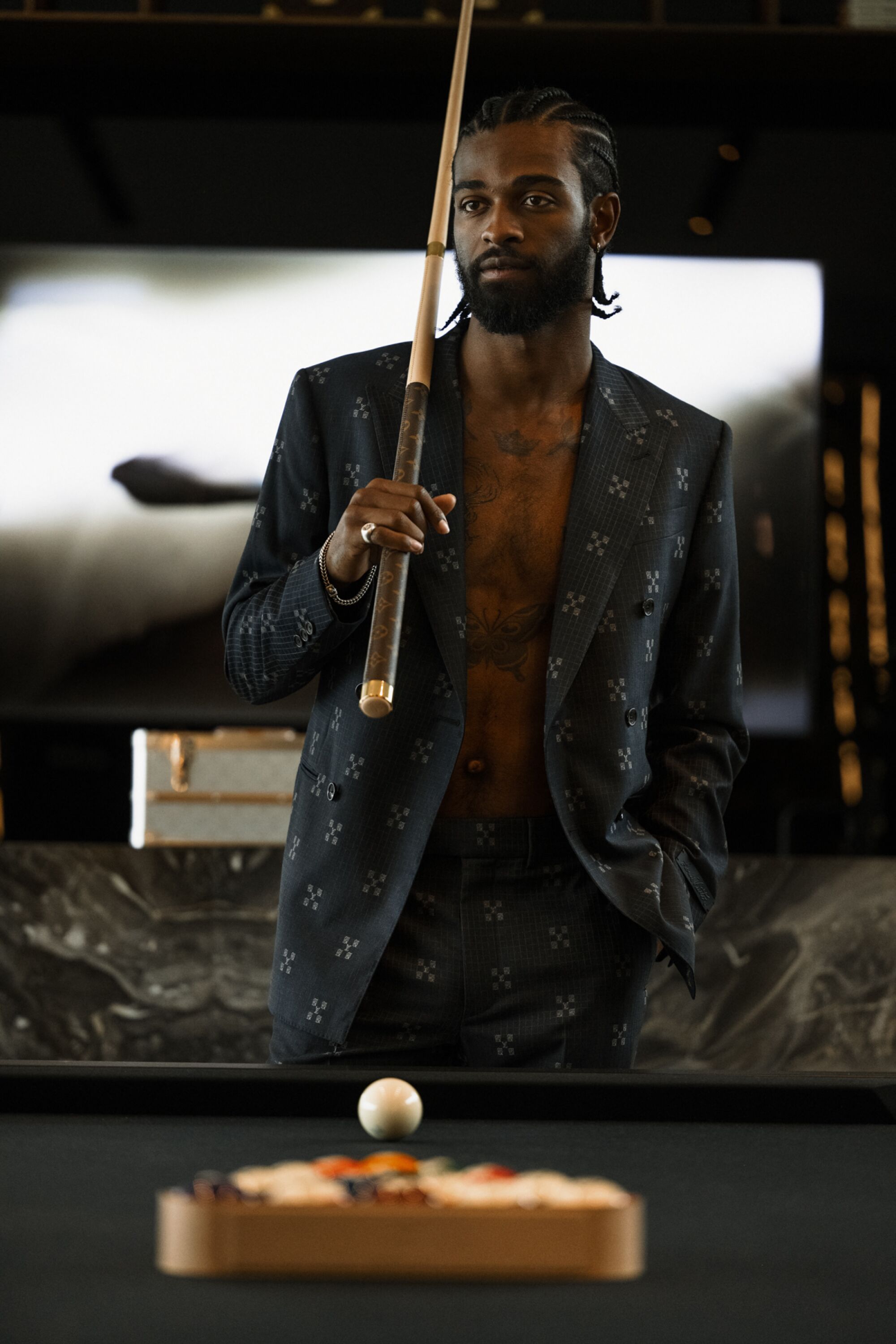 A man in a suit jacket stands in front of a pool table and holds a pool cue.