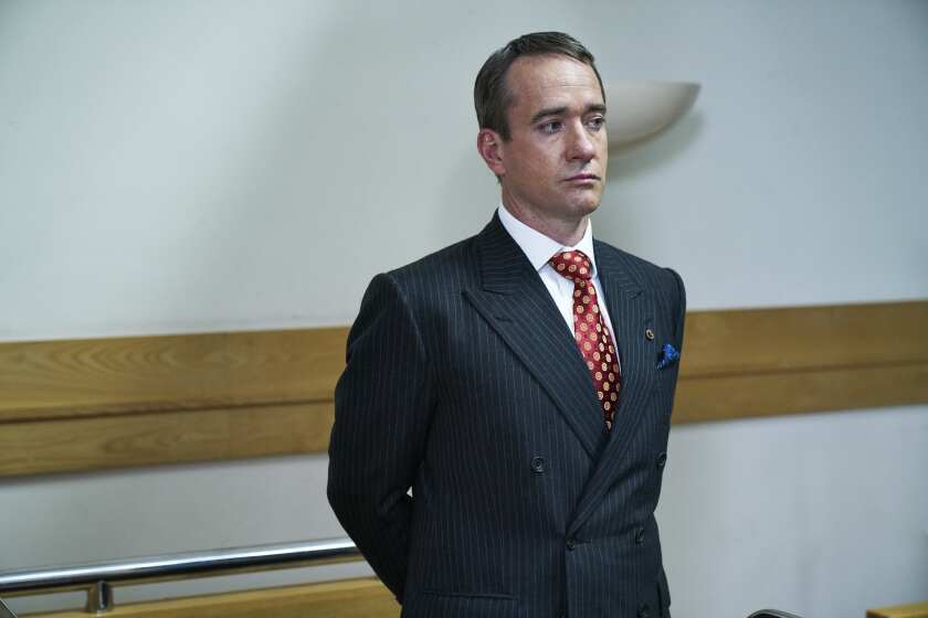 A man in a blue pinstripe suit stands with his arms behind his back.