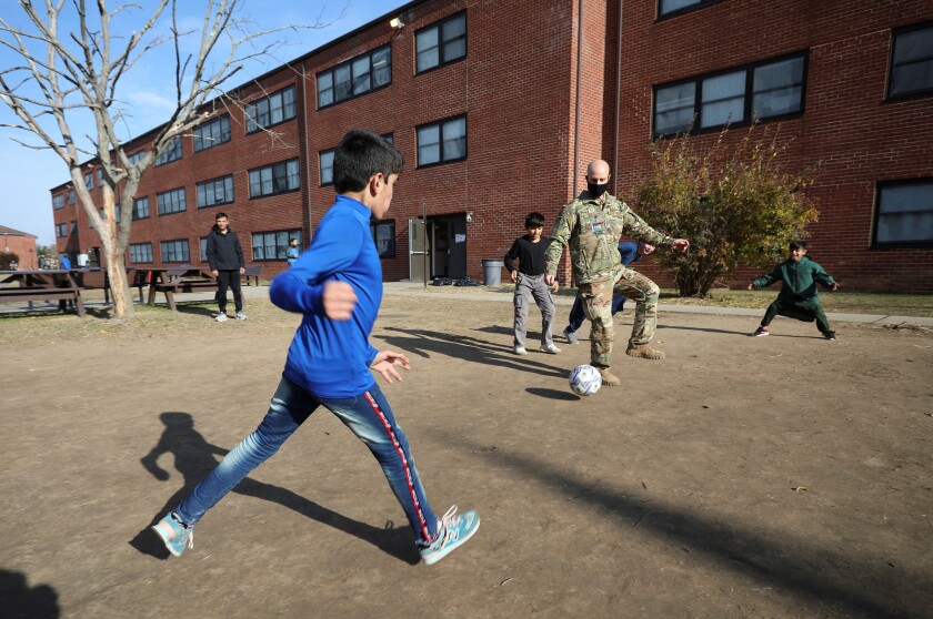 A man in a camouflage uniform plays soccer with children outside a brick building