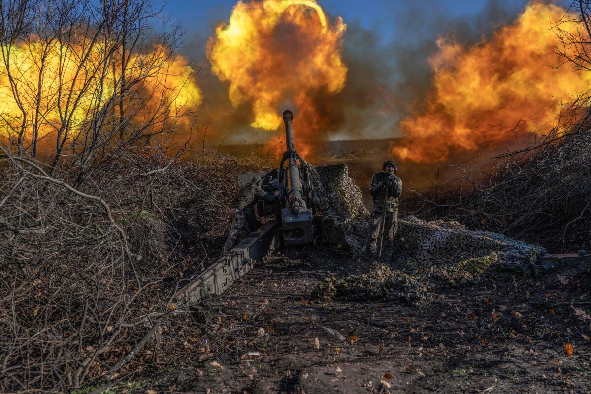 A man stands next to a cannon, with fireballs on the horizon, near another man in a dry brush setting 