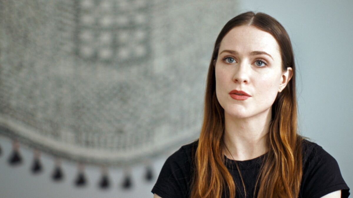 A woman with auburn hair in a black T-shirt speaks to camera