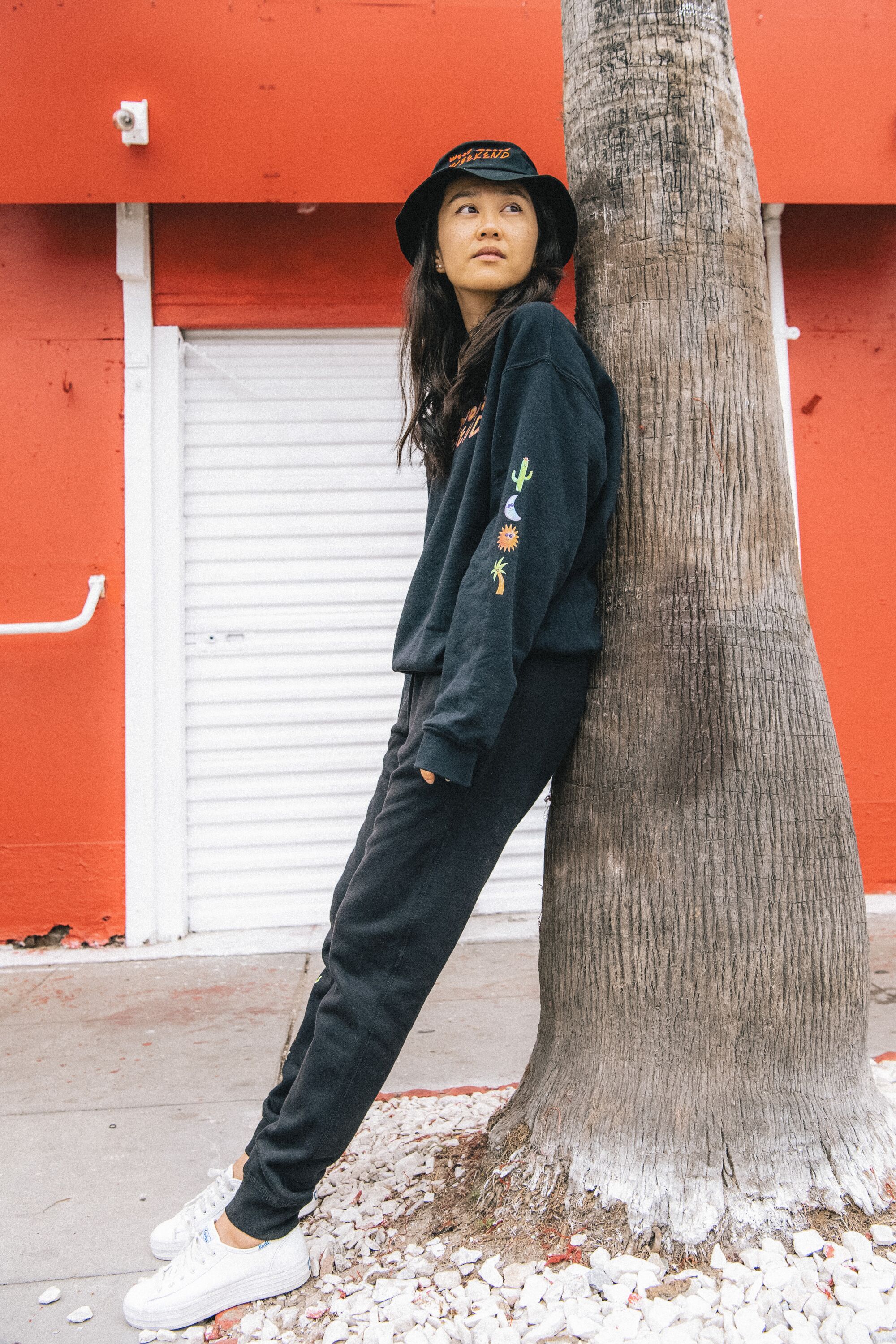 A model in a bucket hat, dark jogging pants and a sweatshirt leans against a tree. 