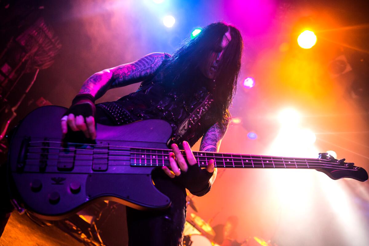 A man with long hair and tattooed arms plays the electric bass on a colorfully lighted concert stage.